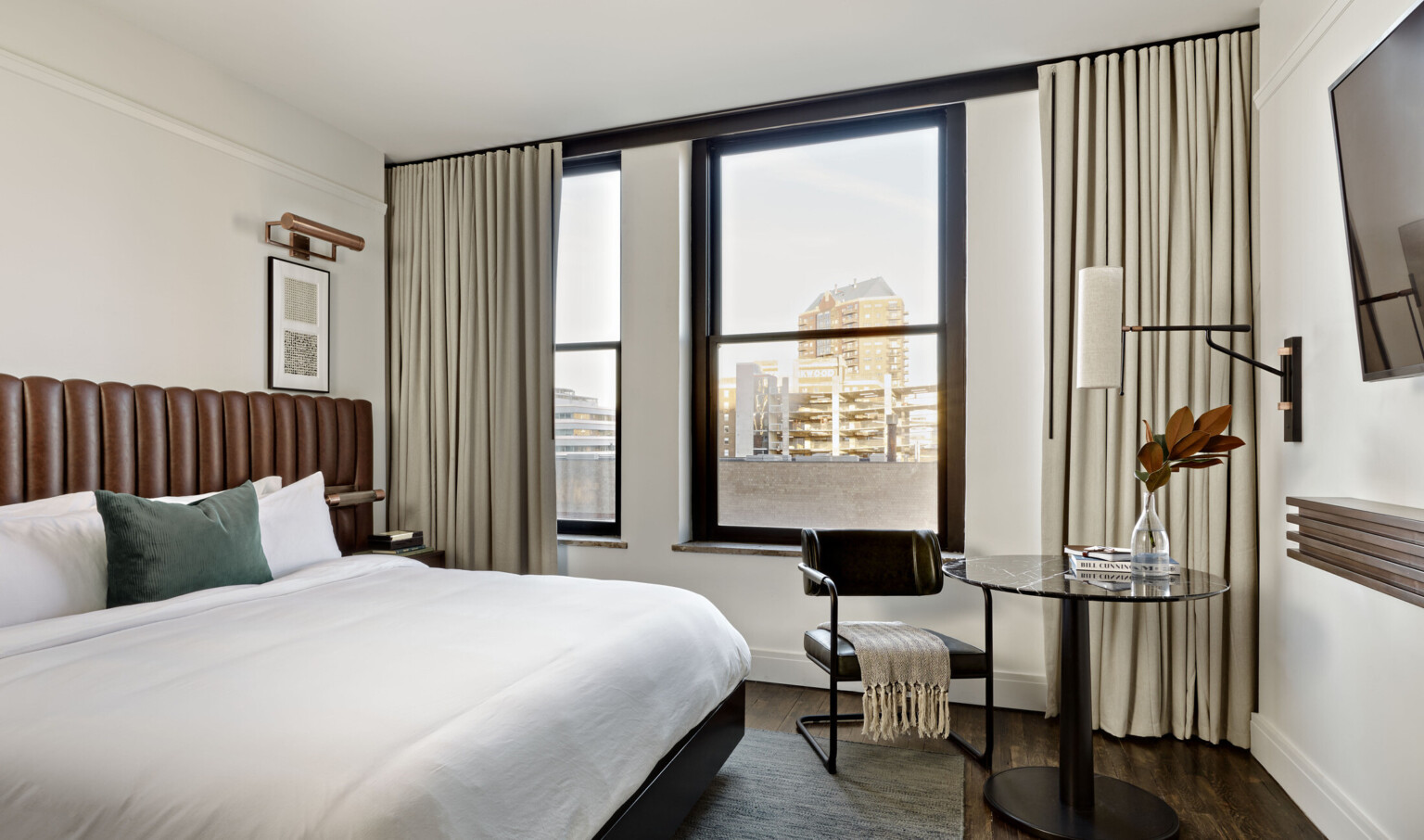 White room with bed with brown headboard, round black table and black cesca chair next to window with view of tall buildings.