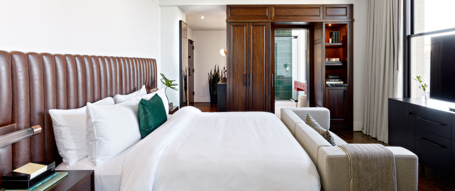 Bedroom with white bed with grey couch at the foot, brown wood built in cupboard and shelves framing pocket door to bathroom