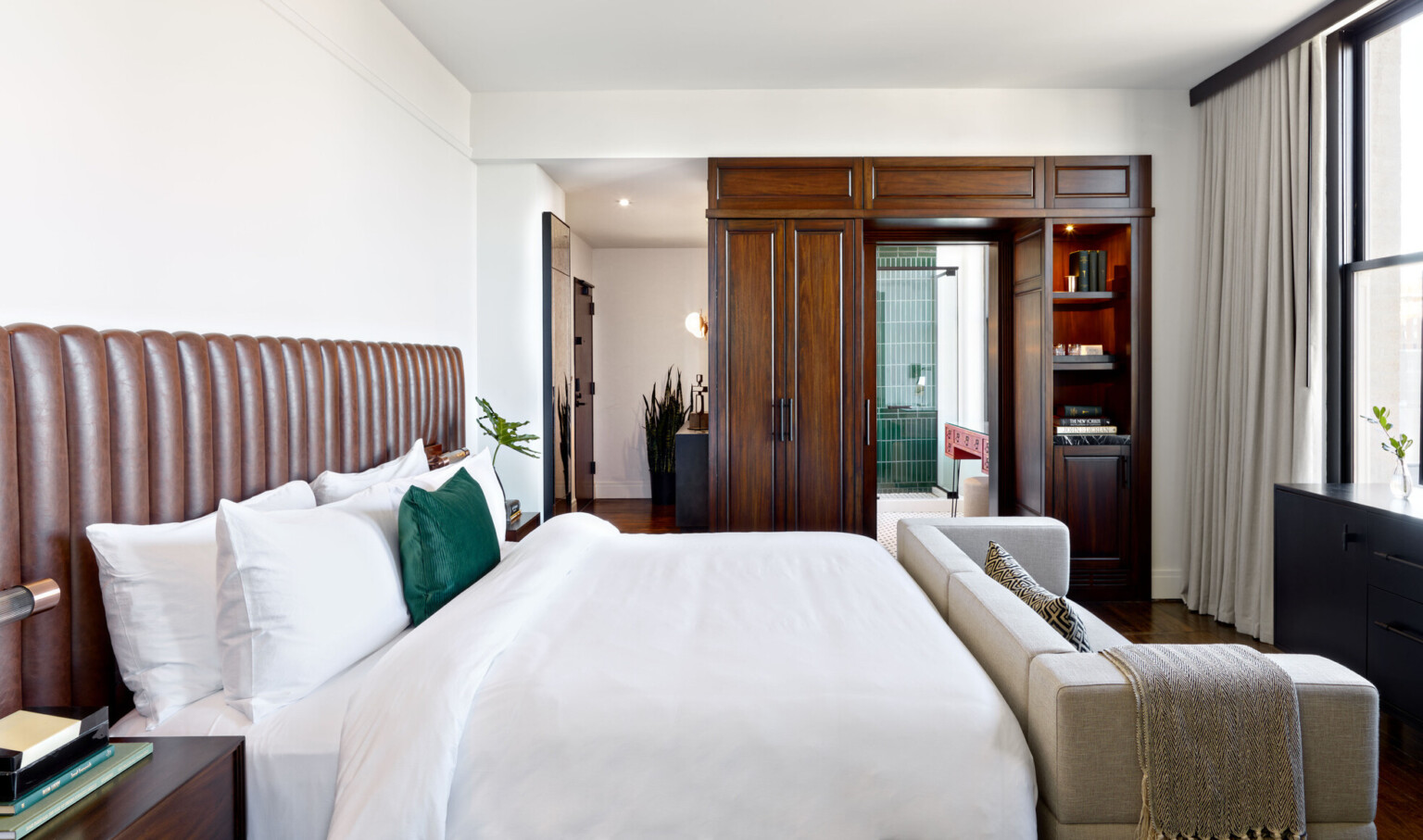 Bedroom with white bed with grey couch at the foot, brown wood built in cupboard and shelves framing pocket door to bathroom
