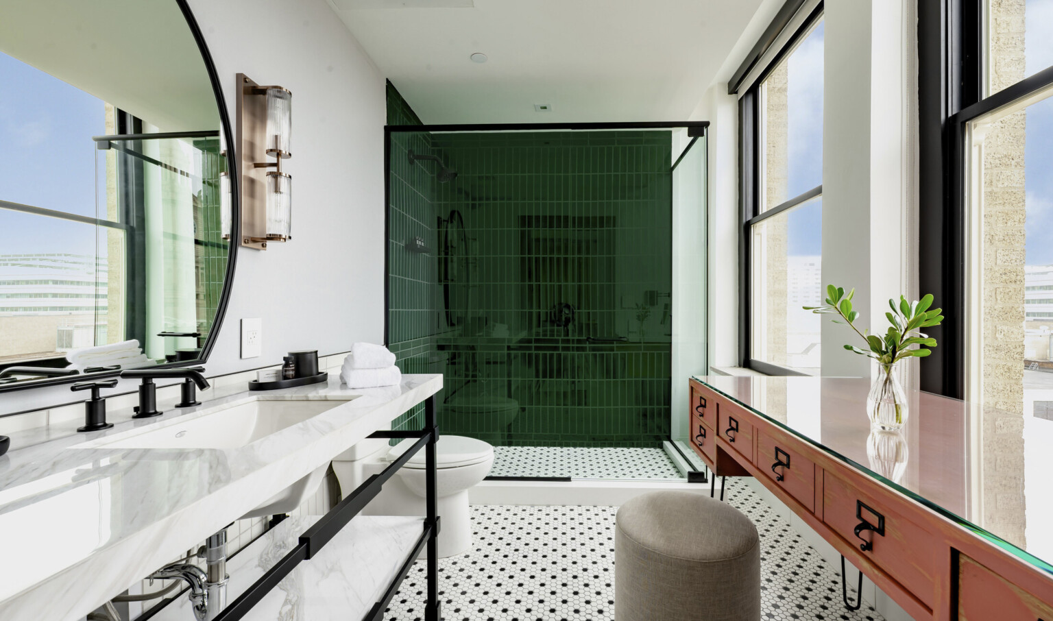 Bathroom with green tiled shower, toilet, and sink left. Right are 2 large windows in front of table and stool