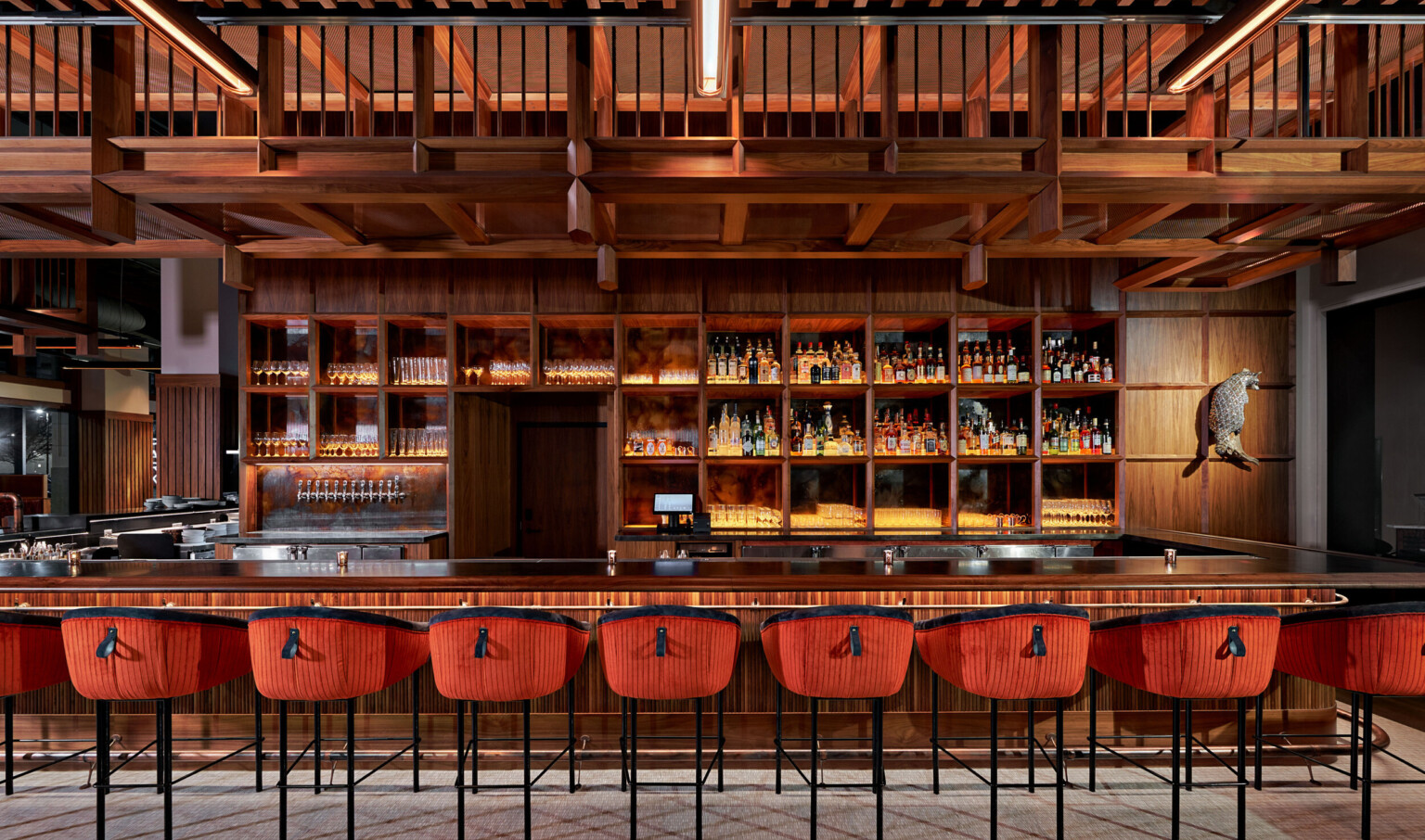 Large wood bar with built in shelves along the back. Wood overhang over back of bar, and stools with red and black upholstery