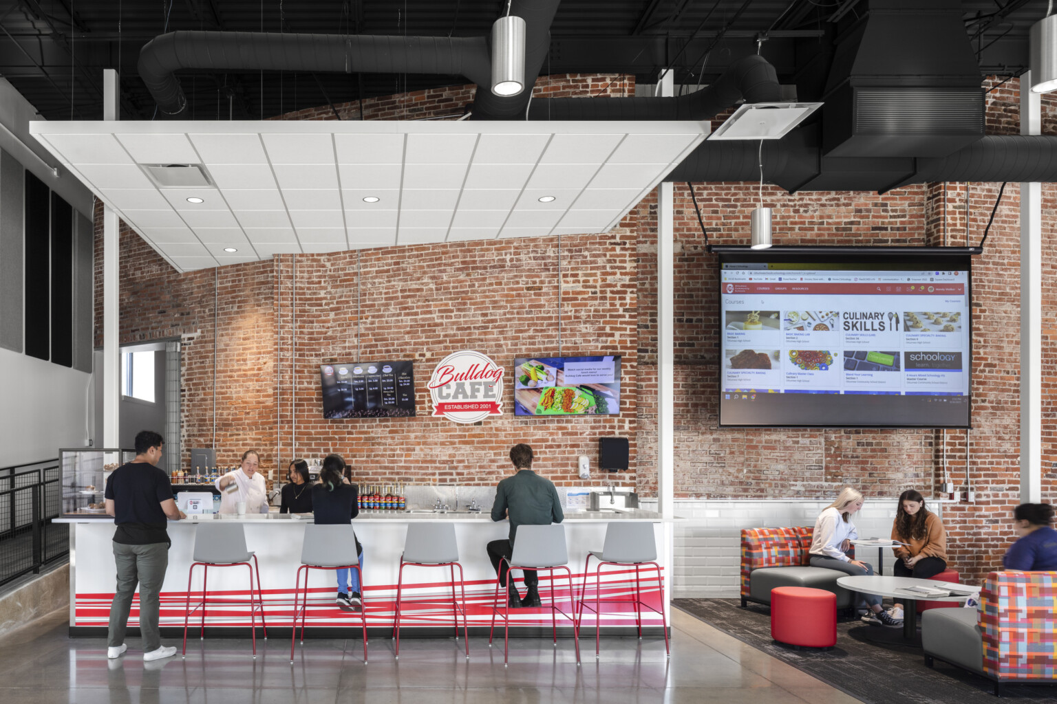 students dine in Bulldog Cafe, exposed brick walls, high-ceilings, exposed ductwork, counter and booth seating, large video screen menu display, polished concrete flooring, signage