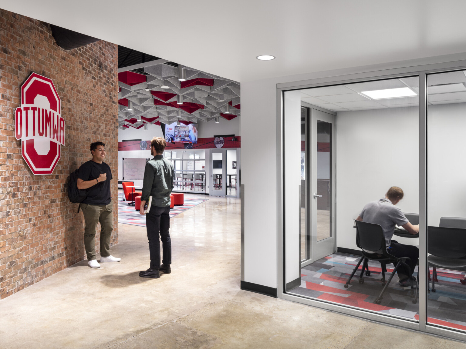 entryway to commons area, signage, two students converse, student in small, glass-walled private work areas provide quiet for focus