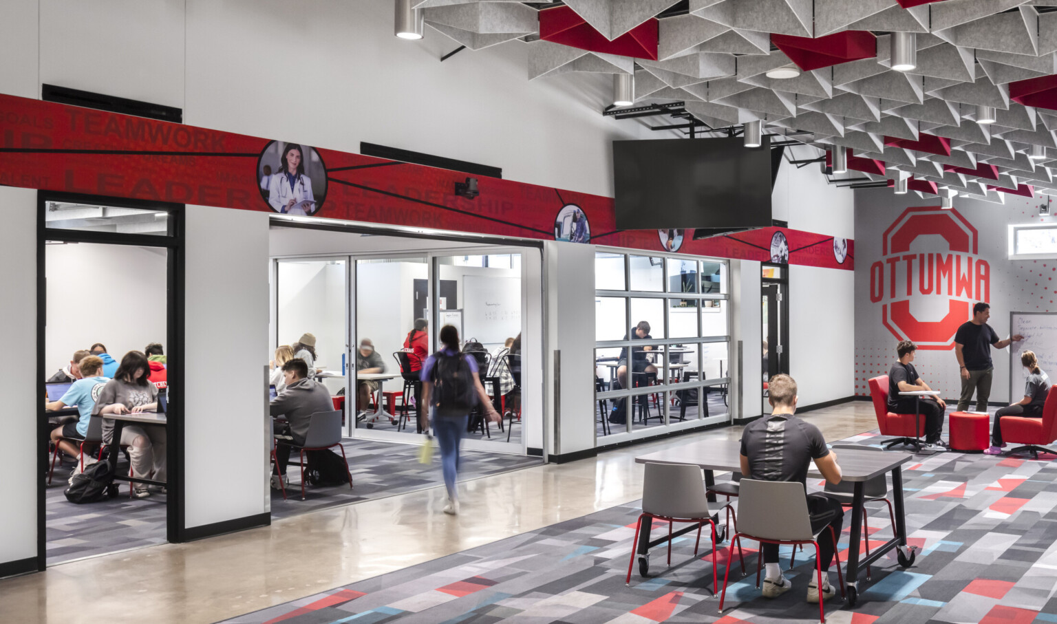 Community space at the Ottumwa Career Campus; red and white branding elements throughout; people studying and engaging throughout the breakout spaces to the left rooms Community space at the Ottumwa Career Campus; red and white branding elements throughout; people studying and engaging throughout the breakout spaces to the left rooms