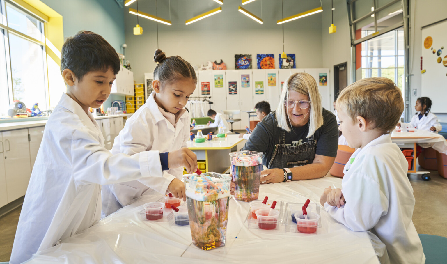 A group of kids in lab coats working with their teacher on a science experience in a lively science classroom