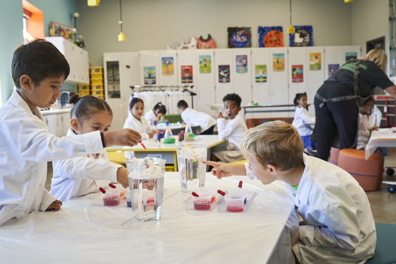 Children in a colorful classroom working on a science experiment with water jugs and pipettes