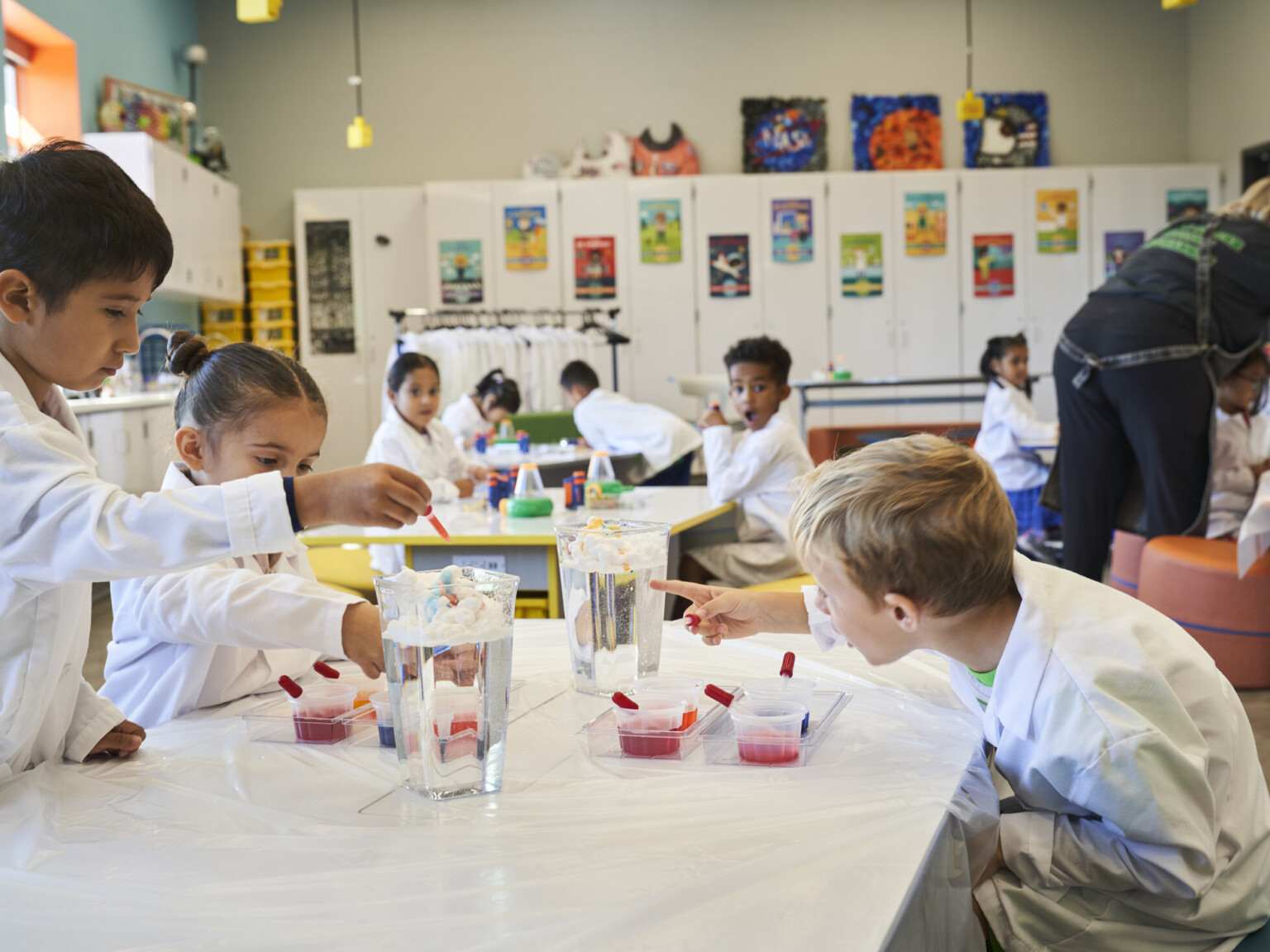 Children in a colorful classroom working on a science experiment with water jugs and pipettes