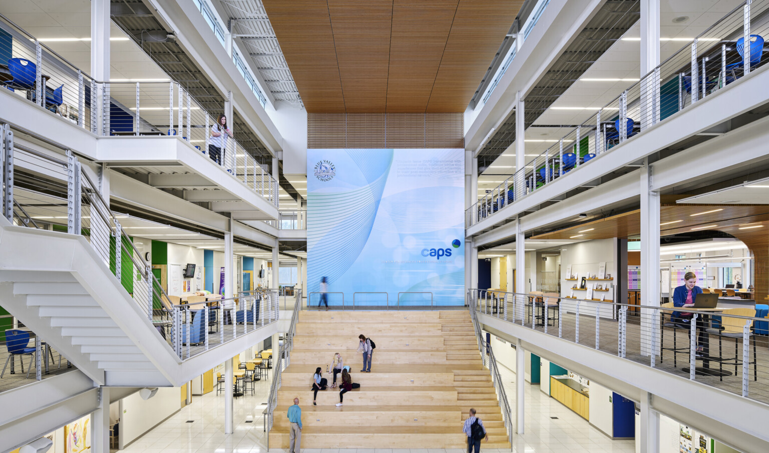 Vibrant atrium with high ceilings, white beams and a large wooden staircase filled with people