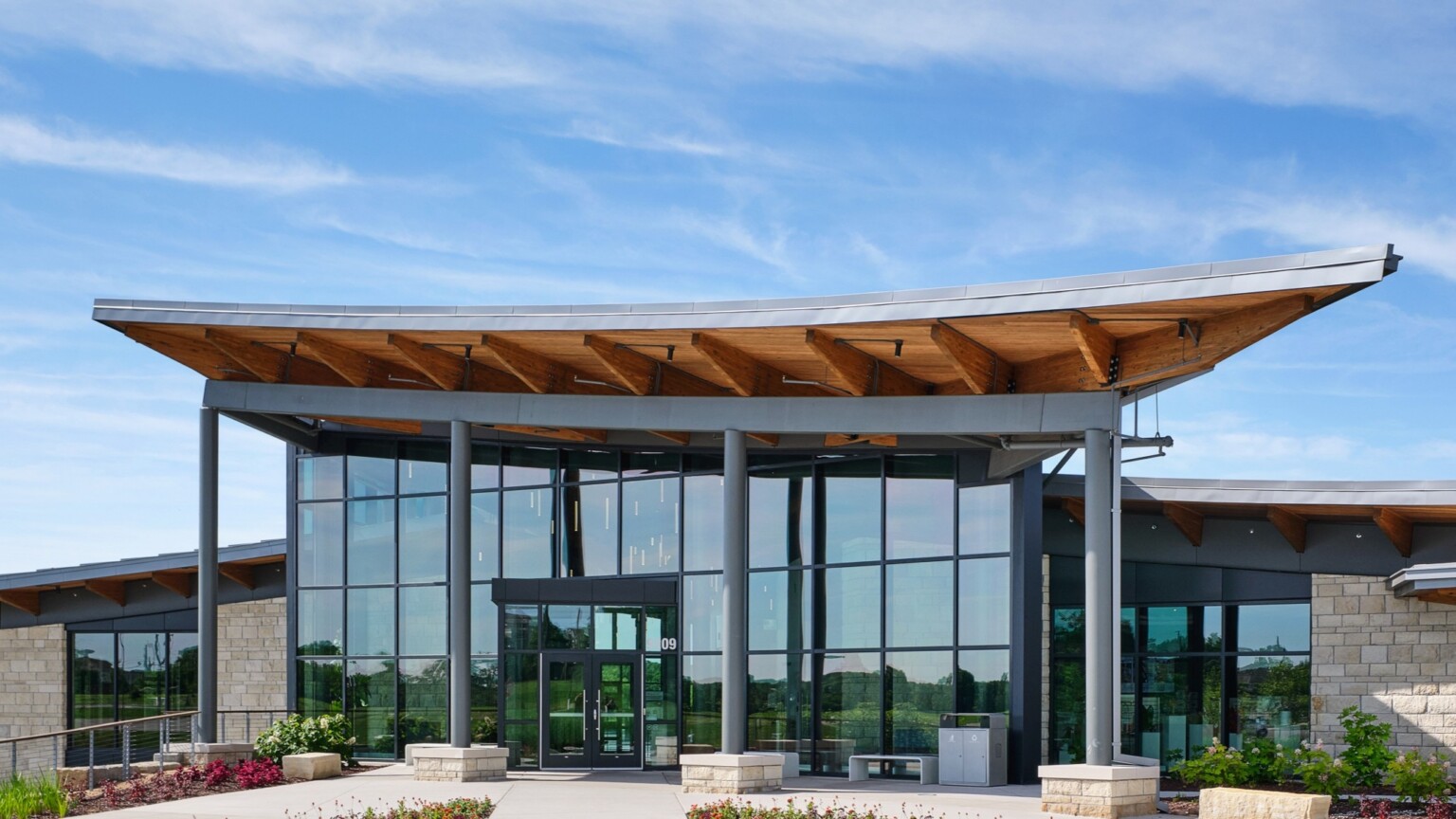 Modern building with floor to ceiling windows surrounding the entrance; large asymmetrical, mass timber awning structure above entrance