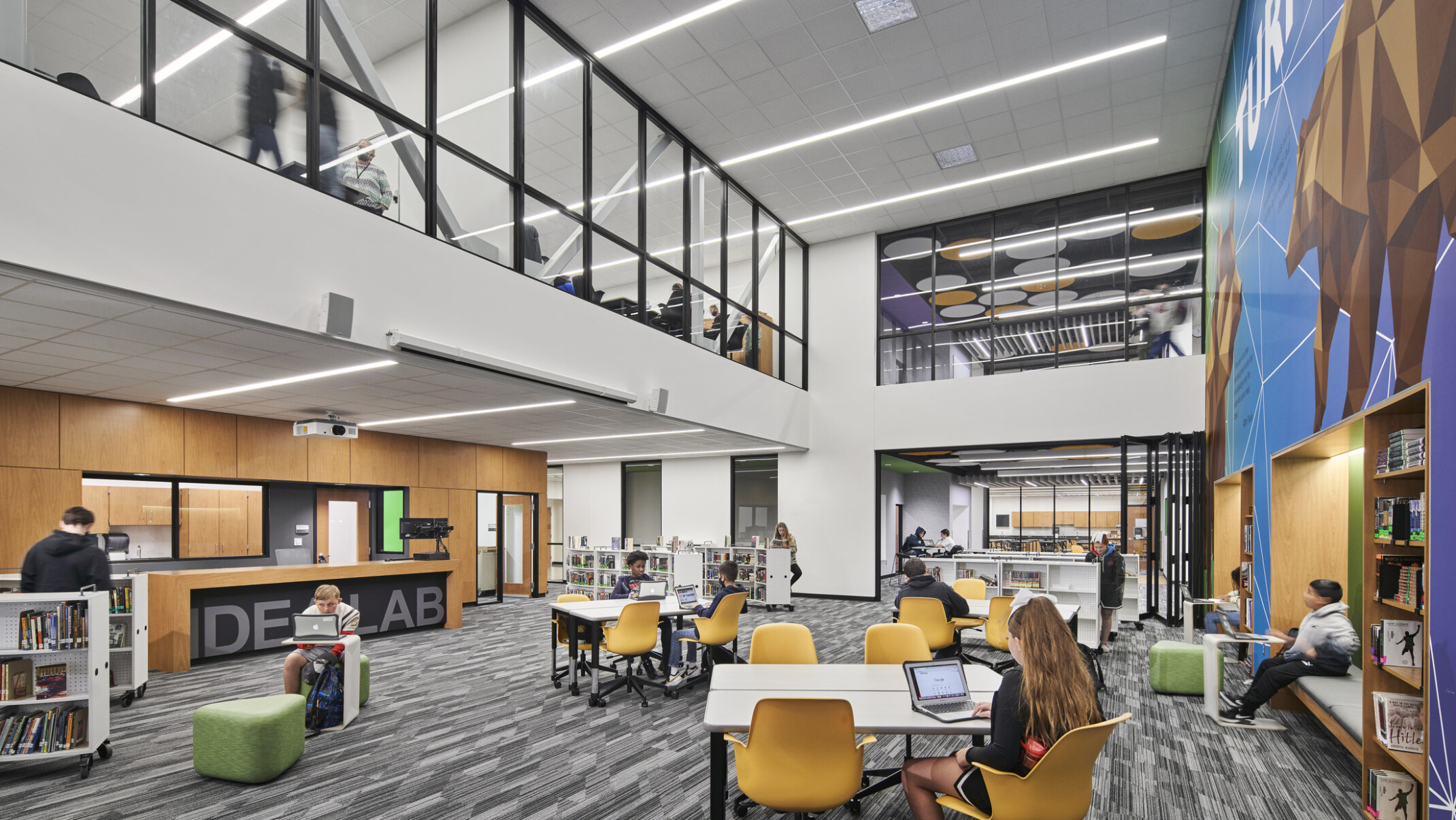 View of main floor of convention center with grey carpet multi-colored wall murals, glass and steel fenestration on second floor media area, various seating groups with tables
