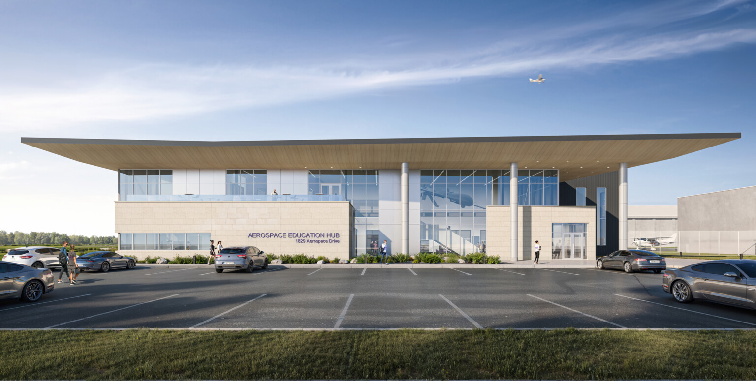 modern two-story building labeled Aerospace Education Hub; sleek design; large glass windows; light-colored stone panels; wide overhanging roof and vertical columns supporting the facade; cars in foreground parking lot; small airplane in the sky above; bright, clear blue sky, light clouds