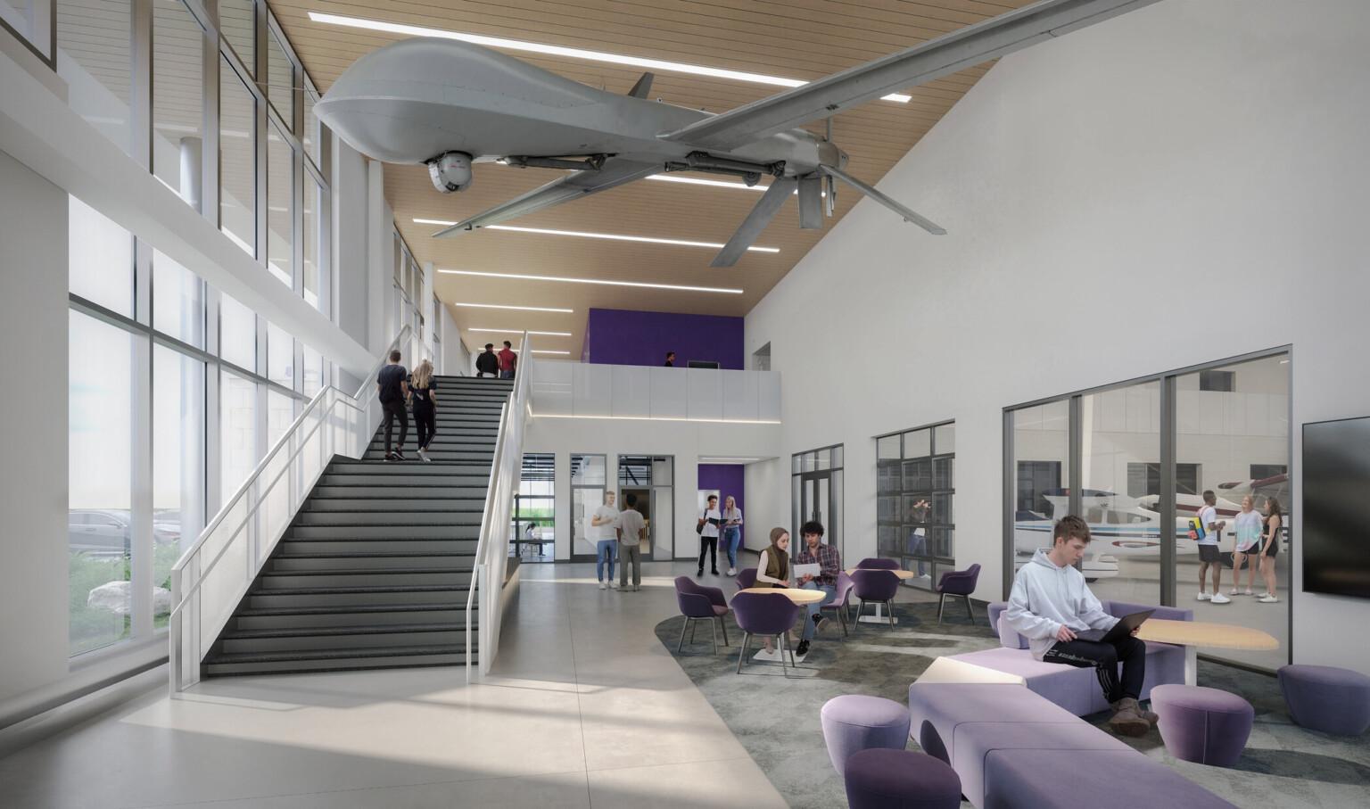 Modern interior of a double height aviation education space; high ceilings and large windows for natural light; Suspended large drone model from ceiling; lower-level tables and chairs for collaborative work; people working on laptops. White walls; gray flooring