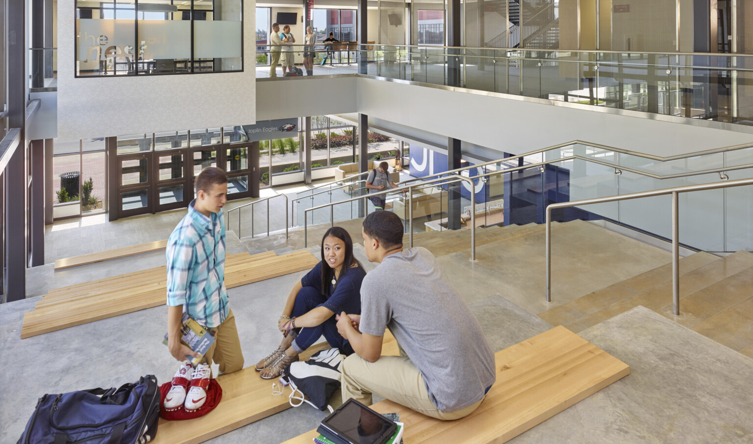 Students sit on concrete stepped seating with wood benches next to stairs, right, leading down to glass double door entry
