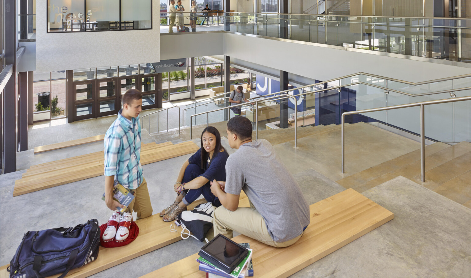 Students sit on concrete stepped seating with wood benches next to stairs, right, leading down to glass double door entry