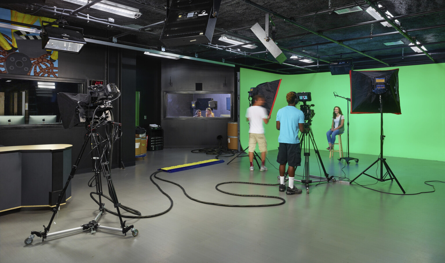 Students operate camera and lighting equipment in front of green screen in black room. Window, left, to editing room