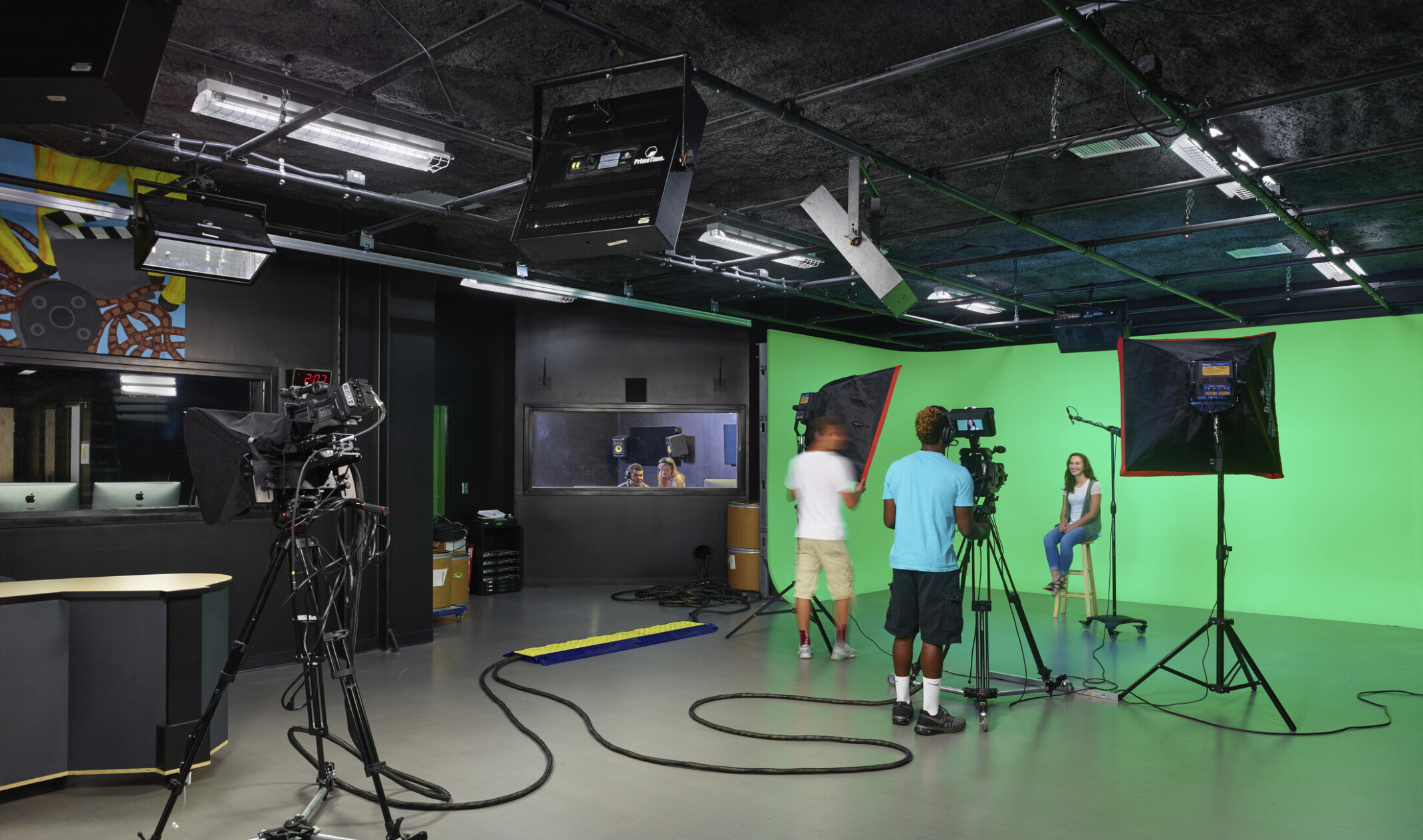 Students operate camera and lighting equipment in front of green screen in black room. Window, left, to editing room