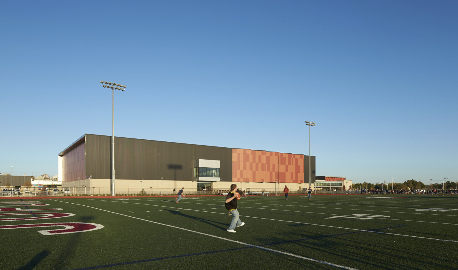 Football field with stadium lights to back of orange panel building with black wrapped facade and orange serrated accents