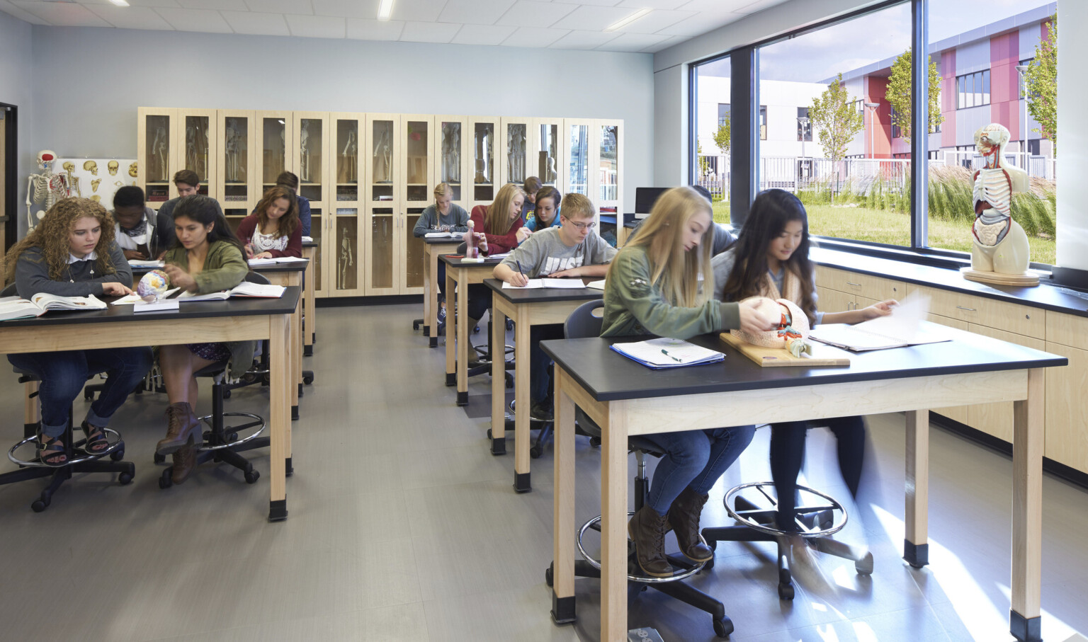 Wood shelving and tables with black counter tops and flexible chairs. Large windows on right classroom wall look to nature