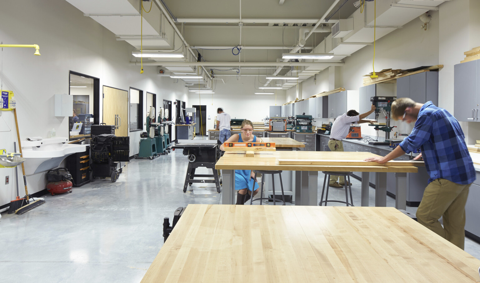 Students work in woodshop, a white room with grey cabinets to right. Power cords hang from ceiling with exposed ducts