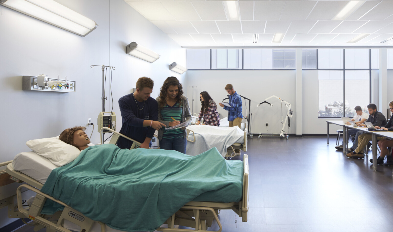 White room with high ceilings and windows on back wall. Hospital beds to left with CPR dummies. Tables face them at right