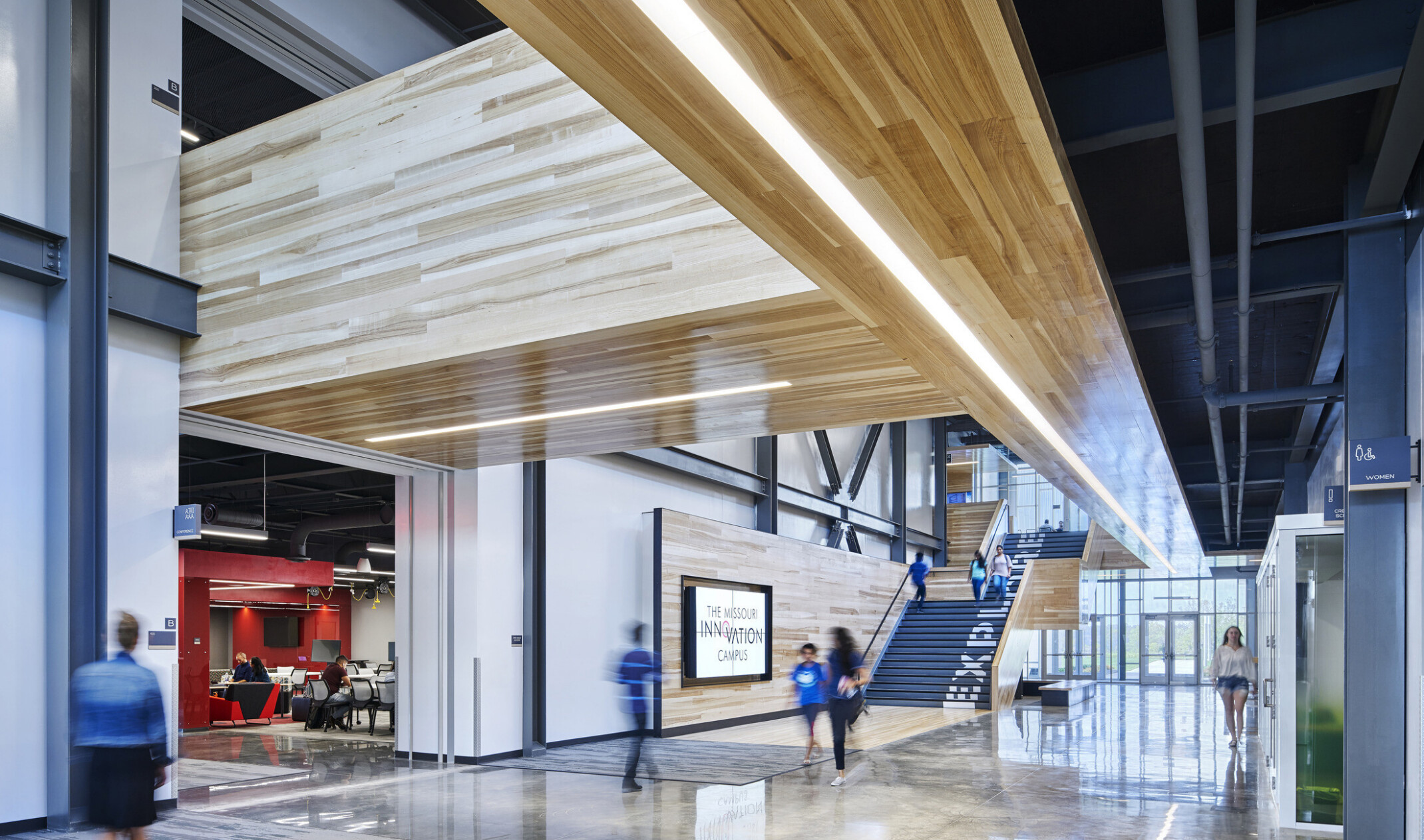 Atrium hallway at entrance with illuminated wood panel walkways above. Black stairs with wood walls has Explore painted up it