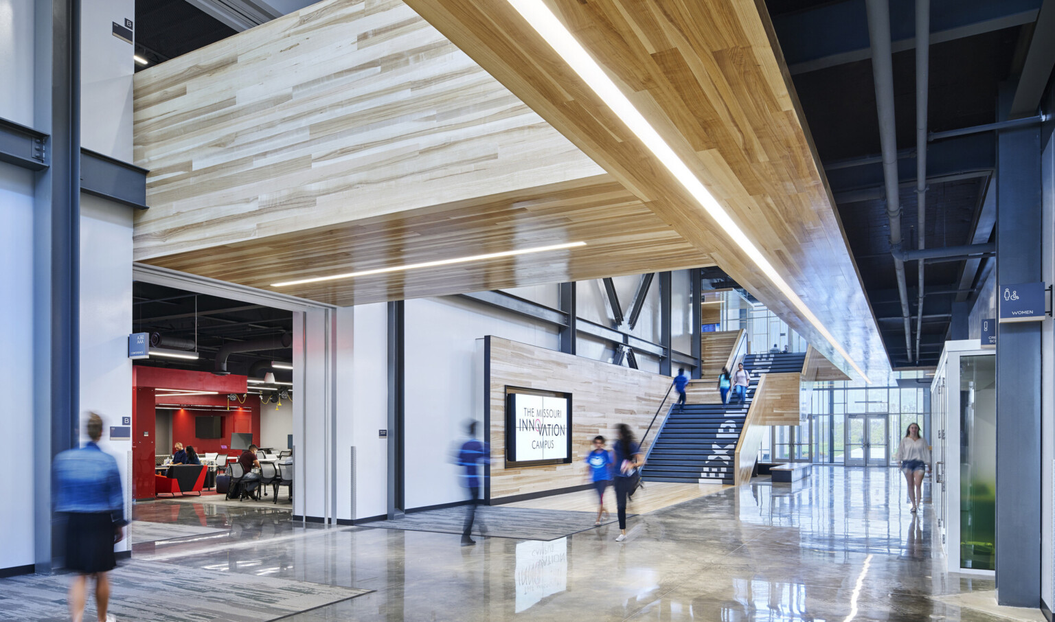 Atrium hallway at entrance with illuminated wood panel walkways above. Black stairs with wood walls has Explore painted up it
