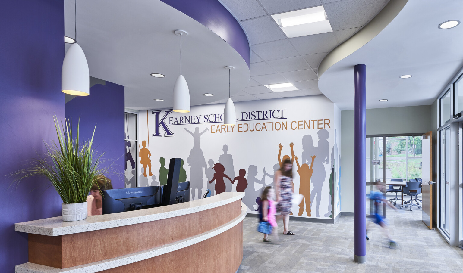 Purple reception area, desk, mural on white wall reading Kearney School District Early Education Center with silhouettes