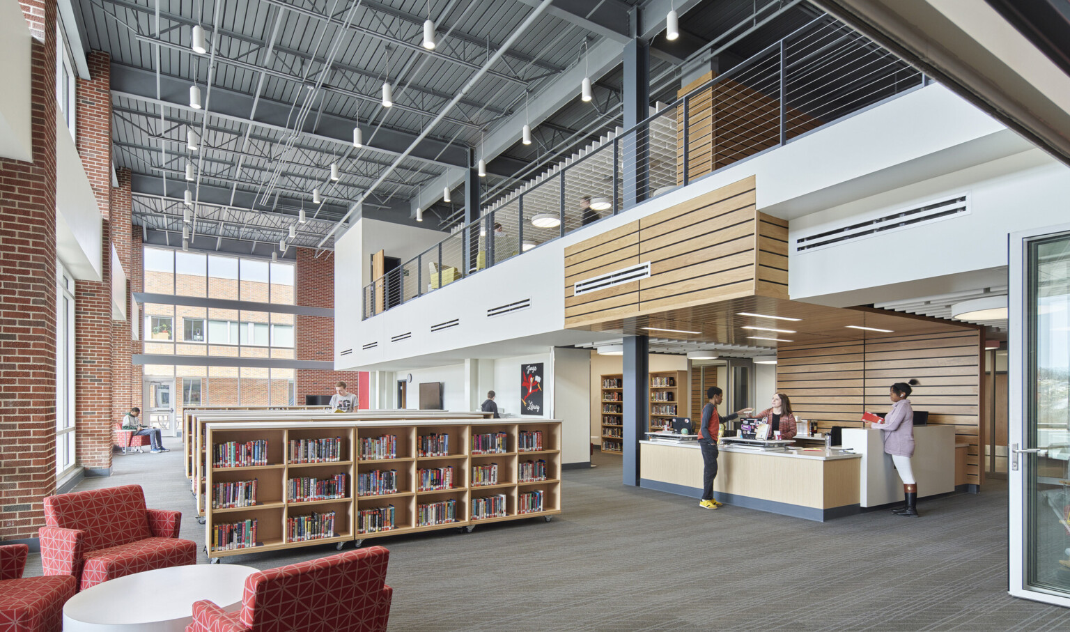 Half height bookcases in library. Double height windows wrapping from left wall to back. Balcony overhang above desk, right