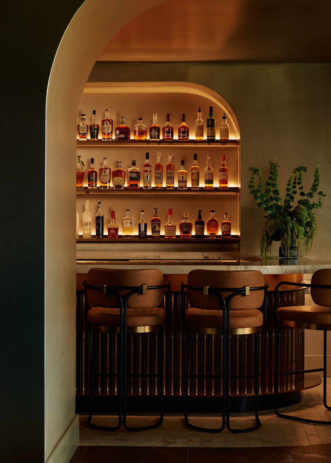 Backlit bar at Momentum KC framed by warm neutral toned arches, featuring illuminated bottle display, fluted bar front detailing, marble countertop, and green floral arrangement