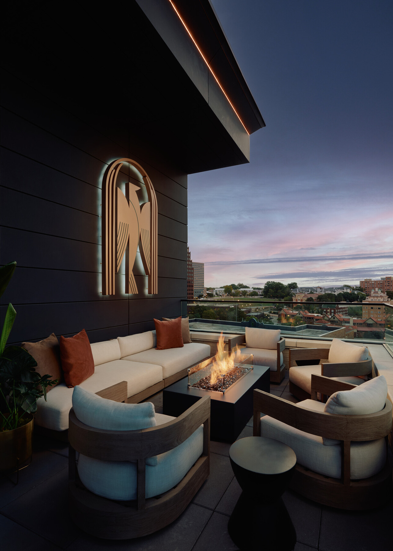 Rooftop terrace at Momentum KC featuring lounge seating arranged around a fire feature at the railing’s edge, with the club’s bird emblem and panoramic evening views beyond.