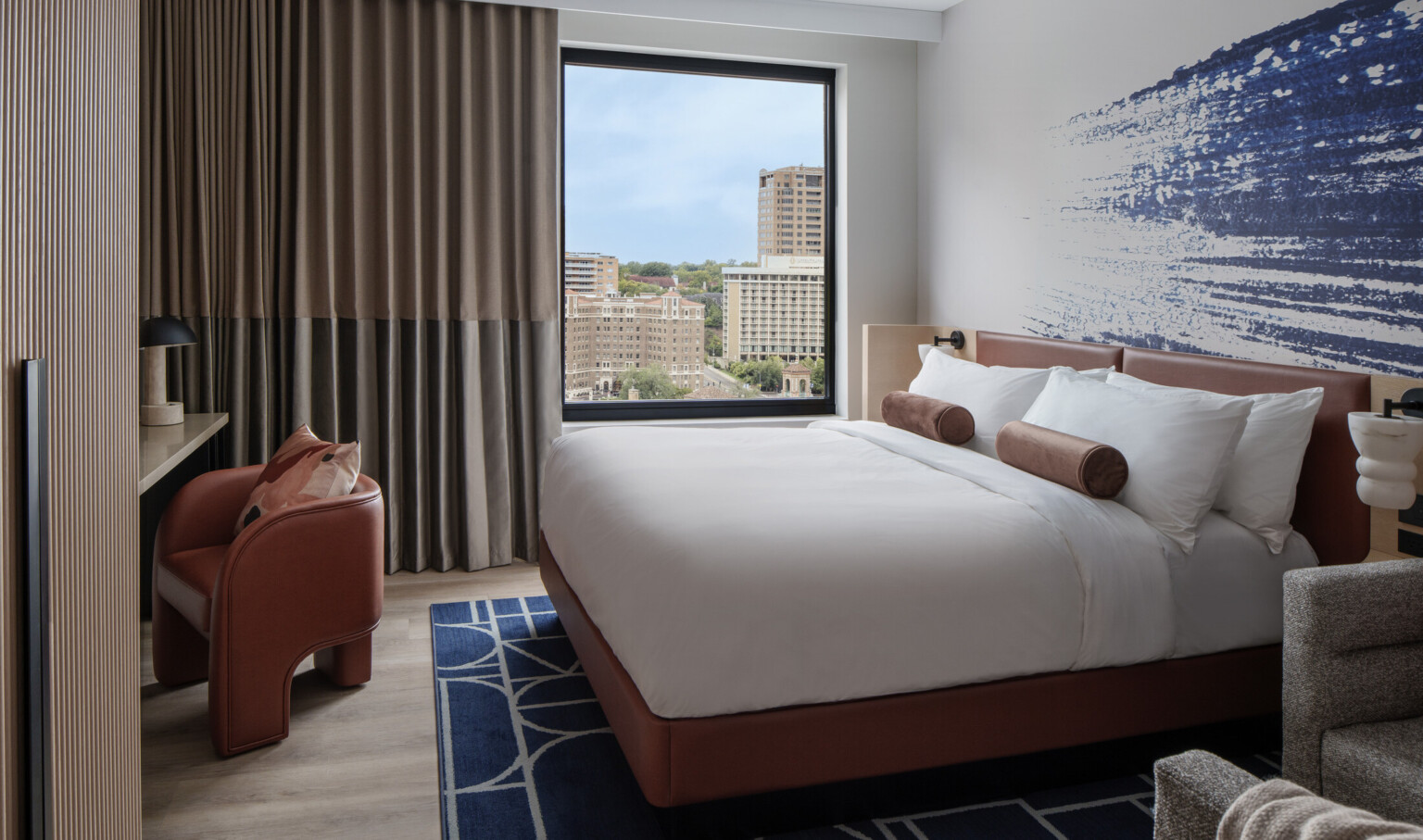 Hotel room with blue artwork behind bed with white linens and dark orange furniture