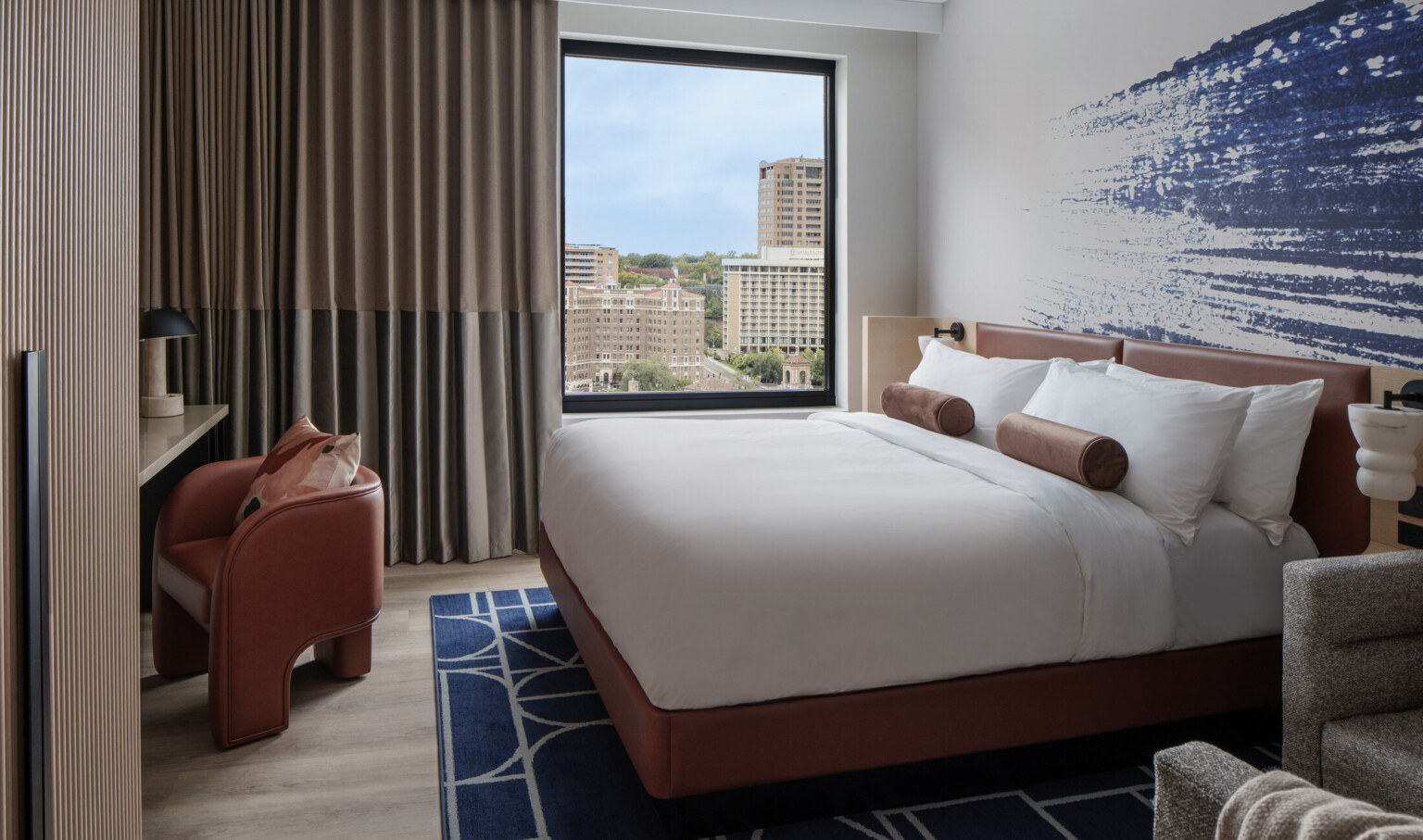Hotel room with blue artwork behind bed with white linens and dark orange furniture