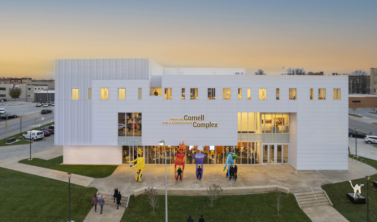 White multistory building light up at dusk by yellow lights; sculptures fill the lawn with people filing in to watch a show at the theater