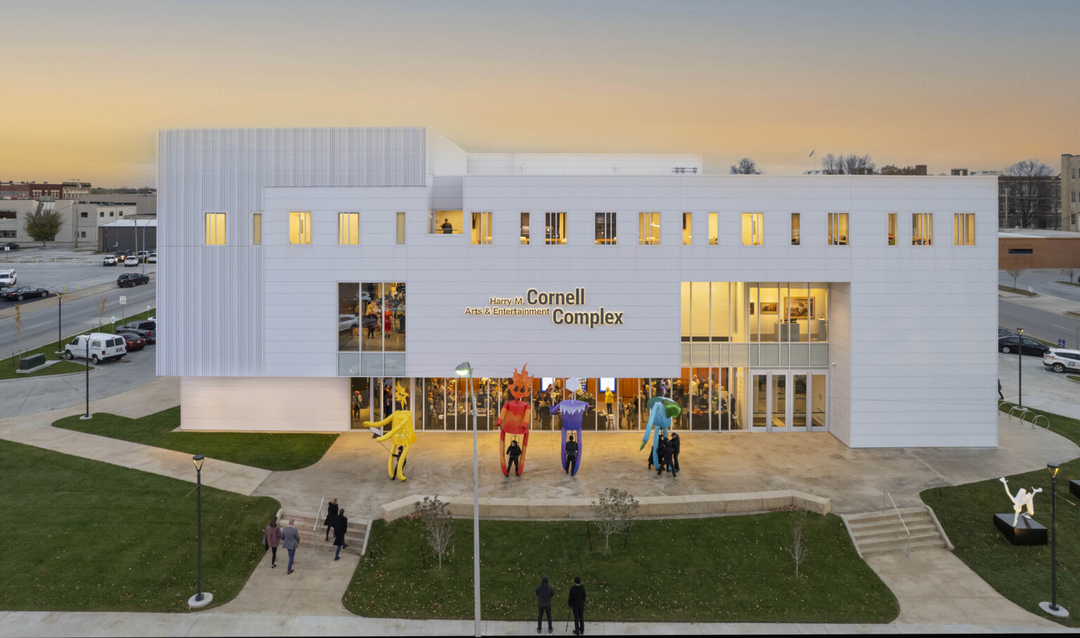 White multistory building light up at dusk by yellow lights; sculptures fill the lawn with people filing in to watch a show at the theater