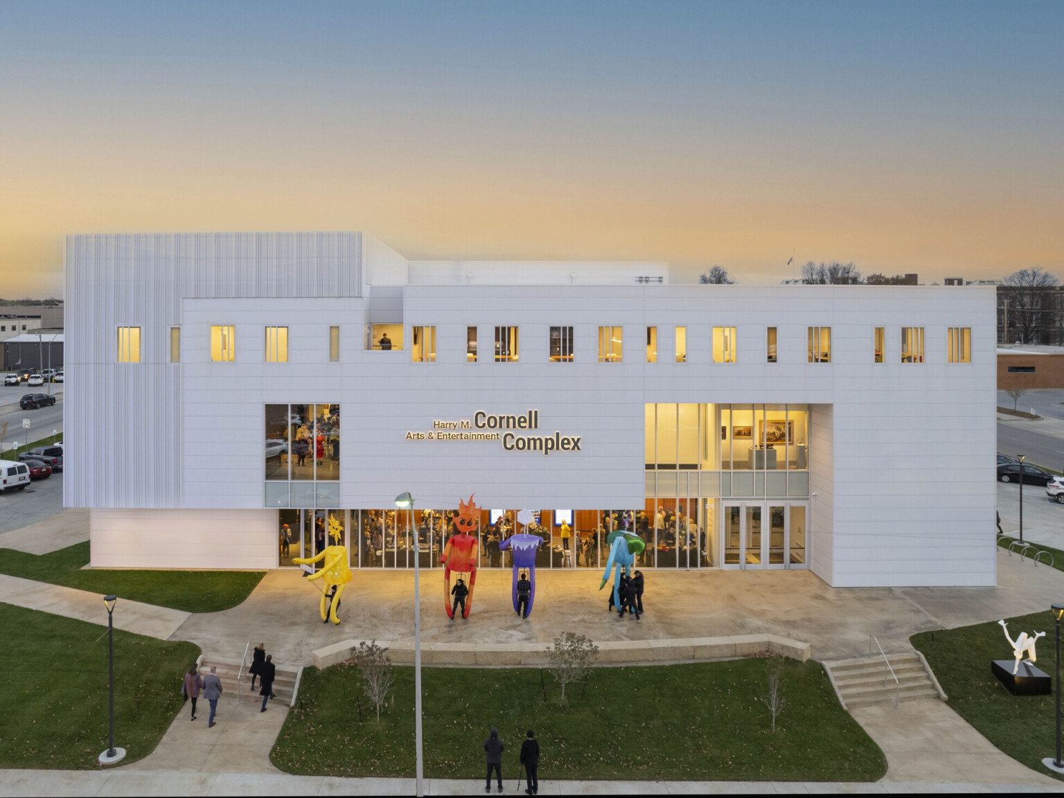 White multistory building light up at dusk by yellow lights; sculptures fill the lawn with people filing in to watch a show at the theater