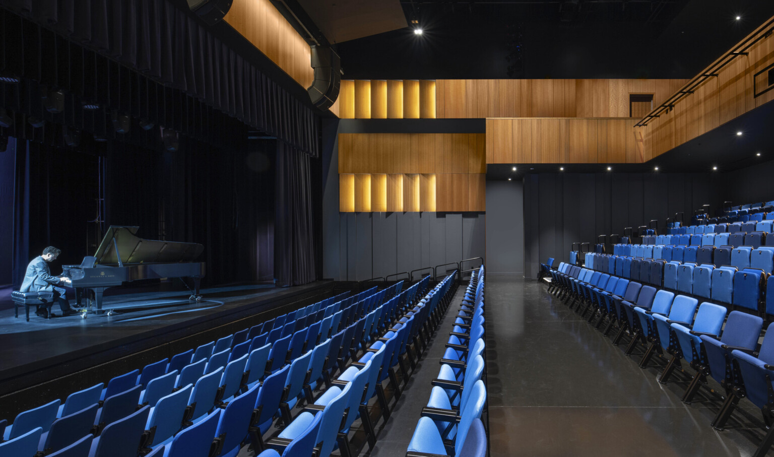 Theater with black walls, wood sections at top and balcony, facing a illuminated stage with pianist playing; blue seating