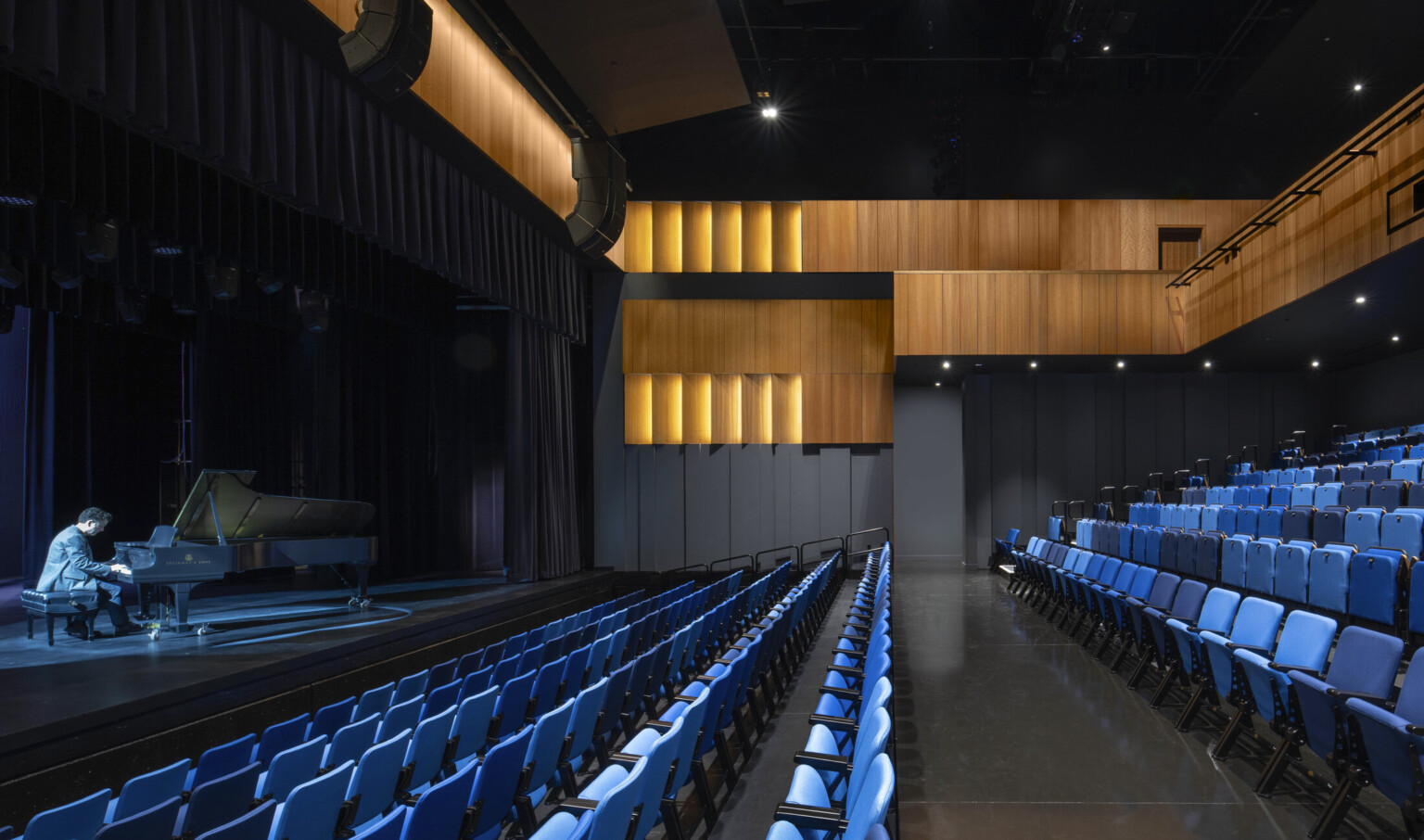 Theater with black walls, wood sections at top and balcony, facing a illuminated stage with pianist playing; blue seating