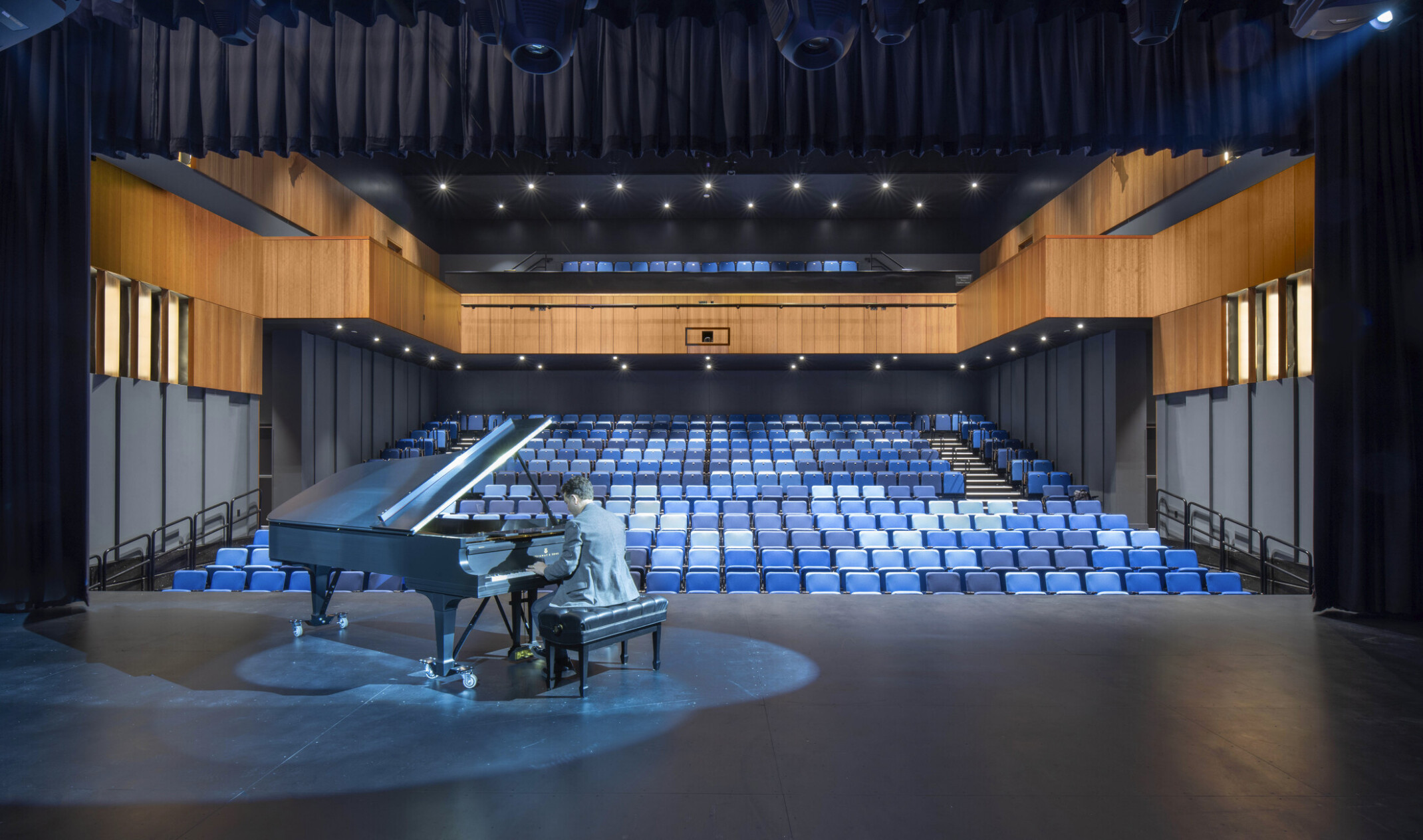 Auditorium theater with piano on stage and pianist performing. Black audience chamber with wood accents and blue seats