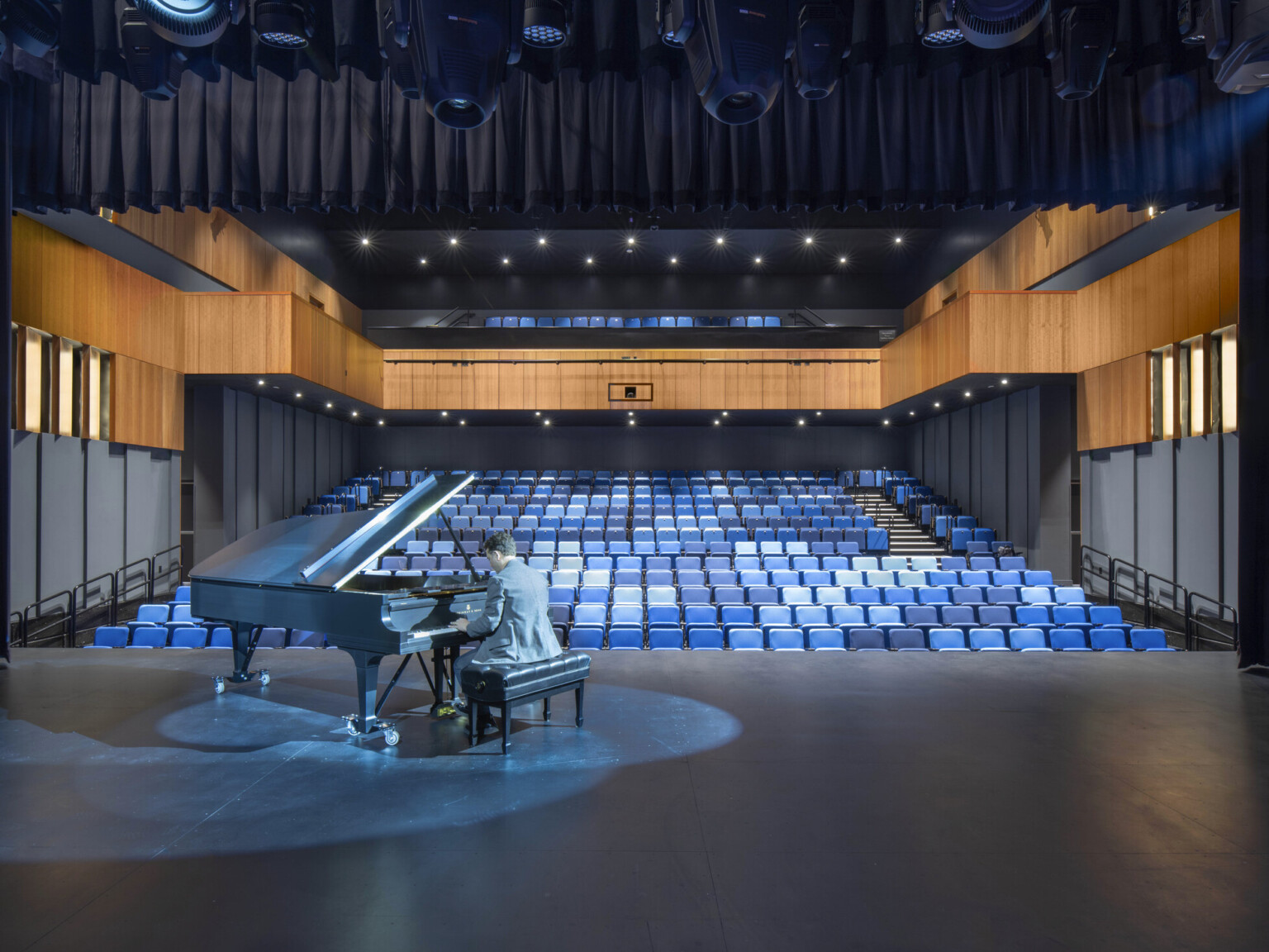 Auditorium theater with piano on stage and pianist performing. Black audience chamber with wood accents and blue seats