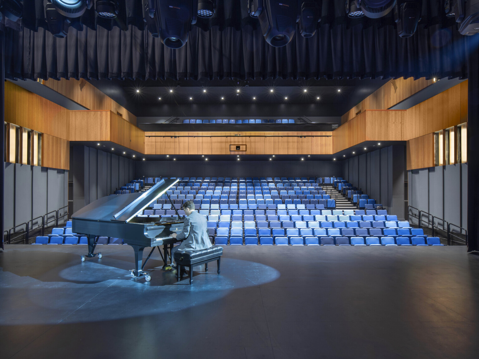 Auditorium theater with piano on stage and pianist performing. Black audience chamber with wood accents and blue seats Auditorium theater with piano on stage and pianist performing. Black audience chamber with wood accents and blue seats