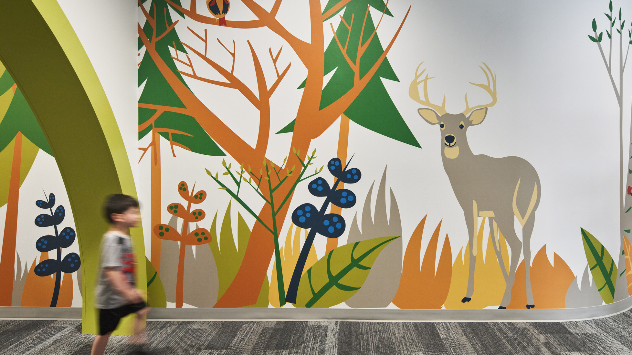 Child walks by colorful mural of cartoon forest and animals in carpeted school hallway, mixed trees, deer, owl, racoon
