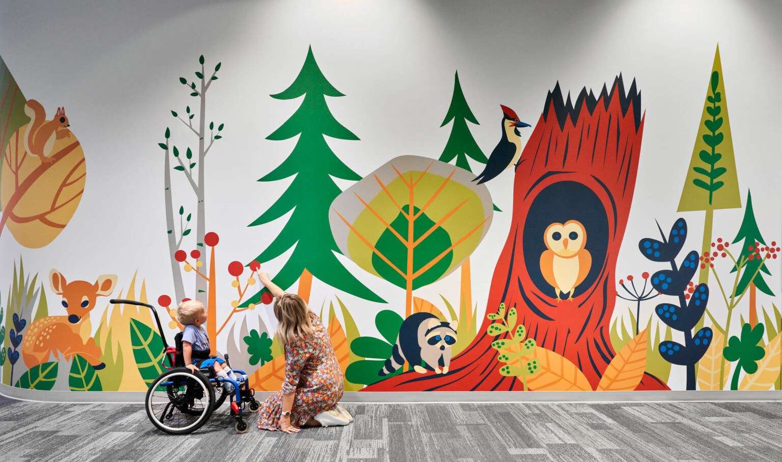 Child and educator interact with mural of cartoon forest and animals in carpeted school hallway. Mixed trees, deer, owl, racoon