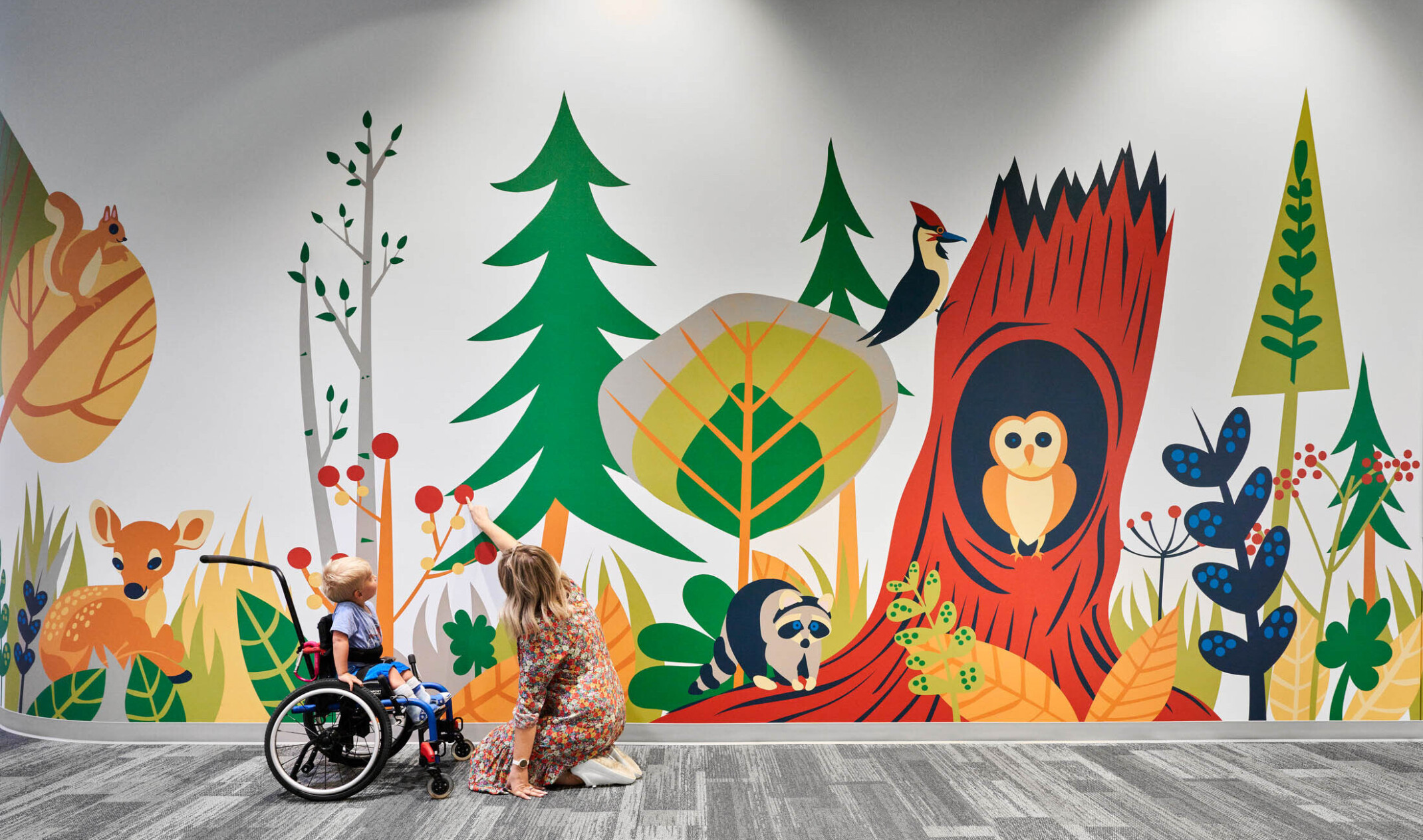 Child and educator interact with mural of cartoon forest and animals in carpeted school hallway. Mixed trees, deer, owl, racoon