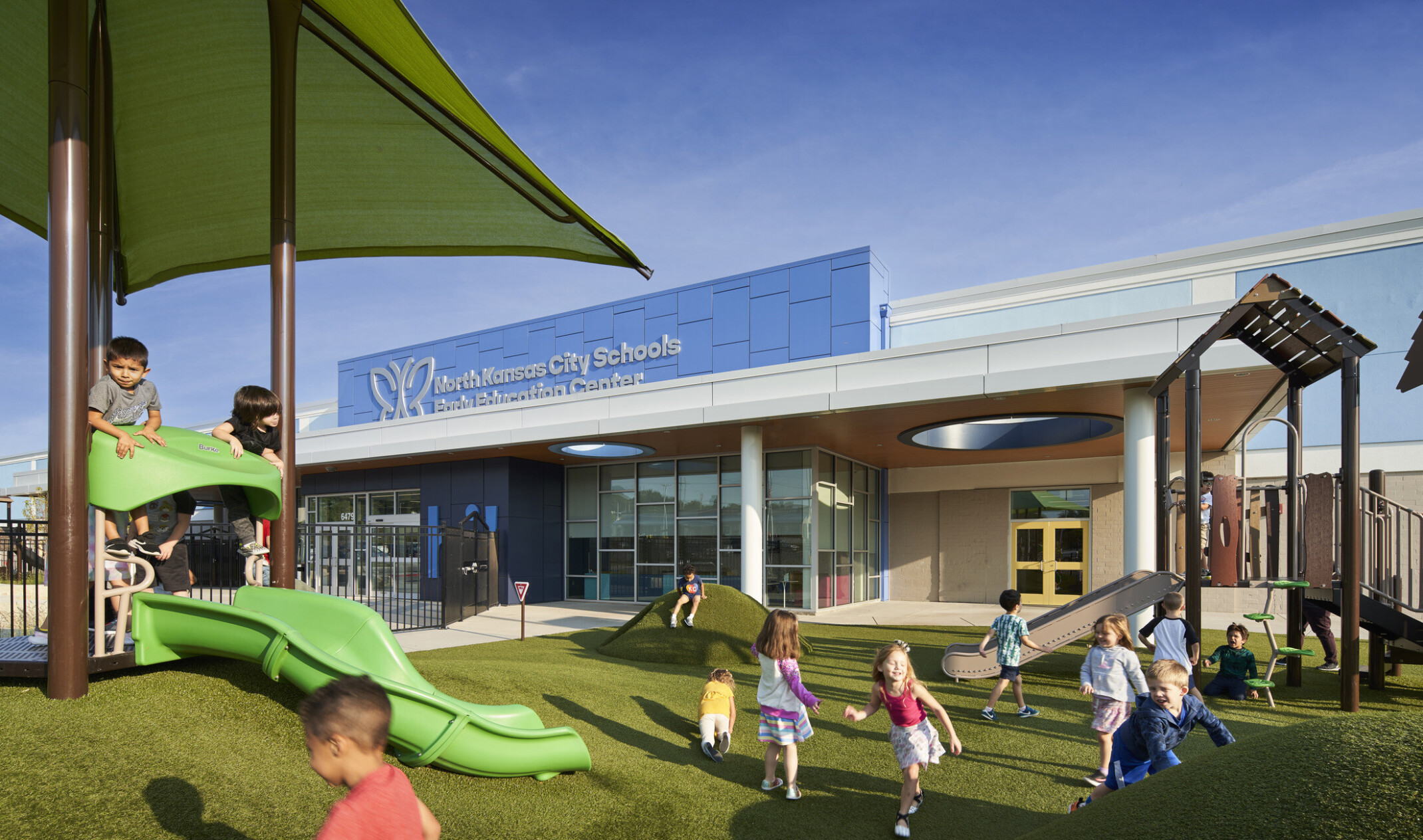 Playground with green turf padding, small hills, slides, and shade near entrance to blue building with North Kansas City Early Education Center sign