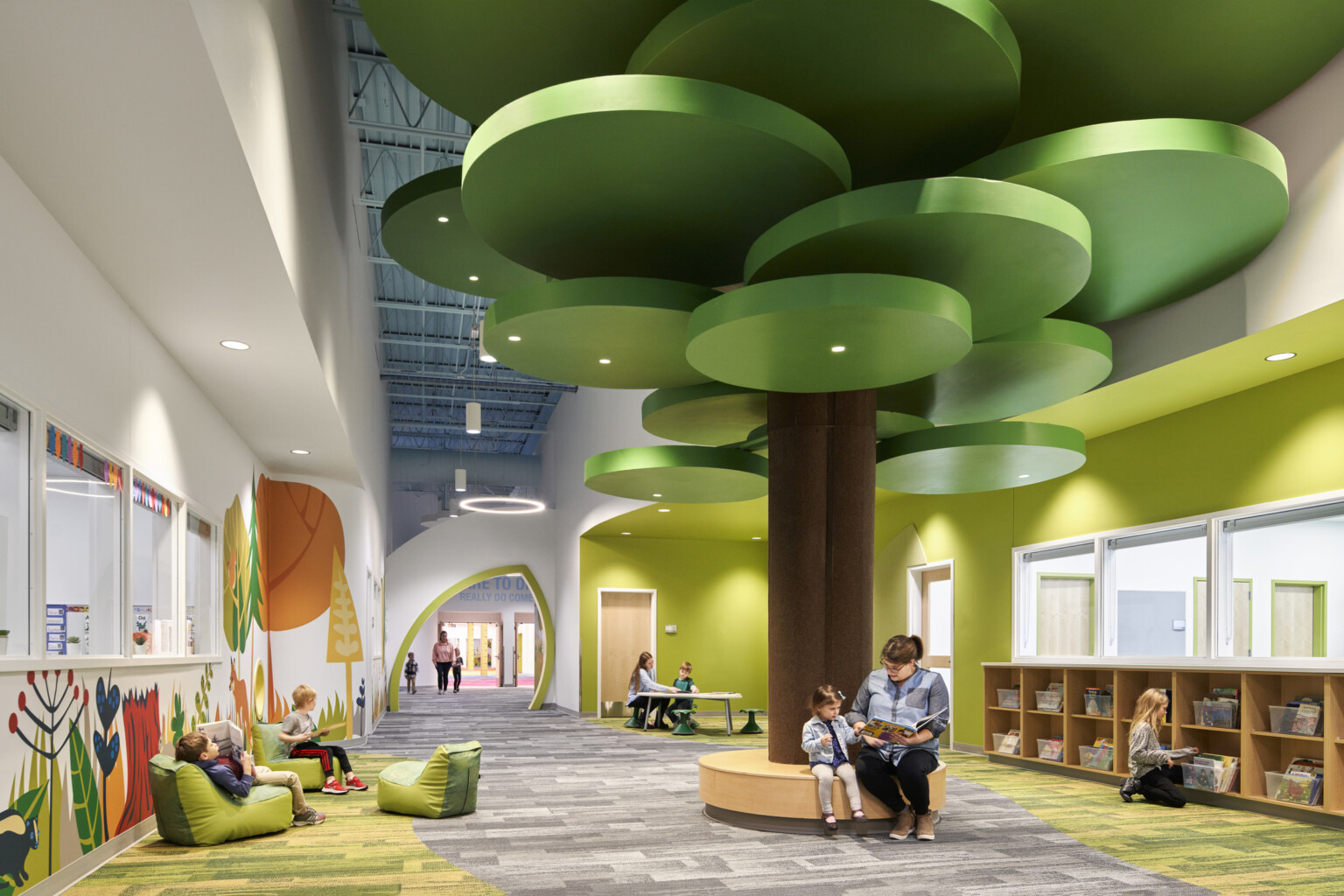 people reading on a brown circular bench with a brown column and green ceiling fixtures in hallway lined with forest graphics; dlr group design people reading on a brown circular bench with a brown column and green ceiling fixtures in hallway lined with forest graphics; dlr group design