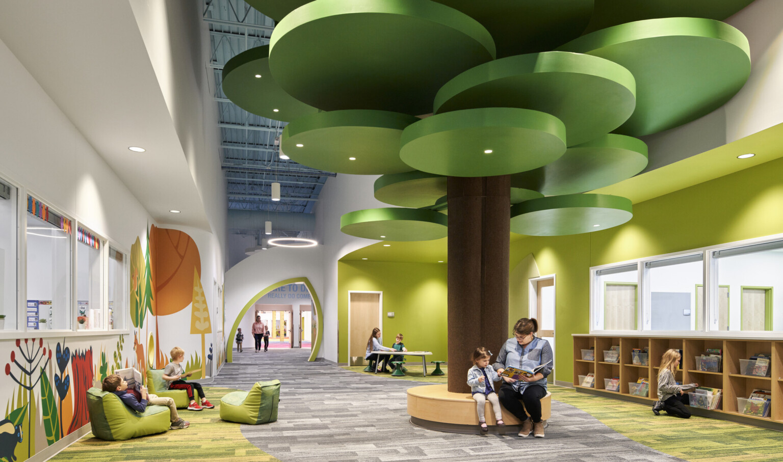 people reading on a brown circular bench with a brown column and green ceiling fixtures in hallway lined with forest graphics; dlr group design