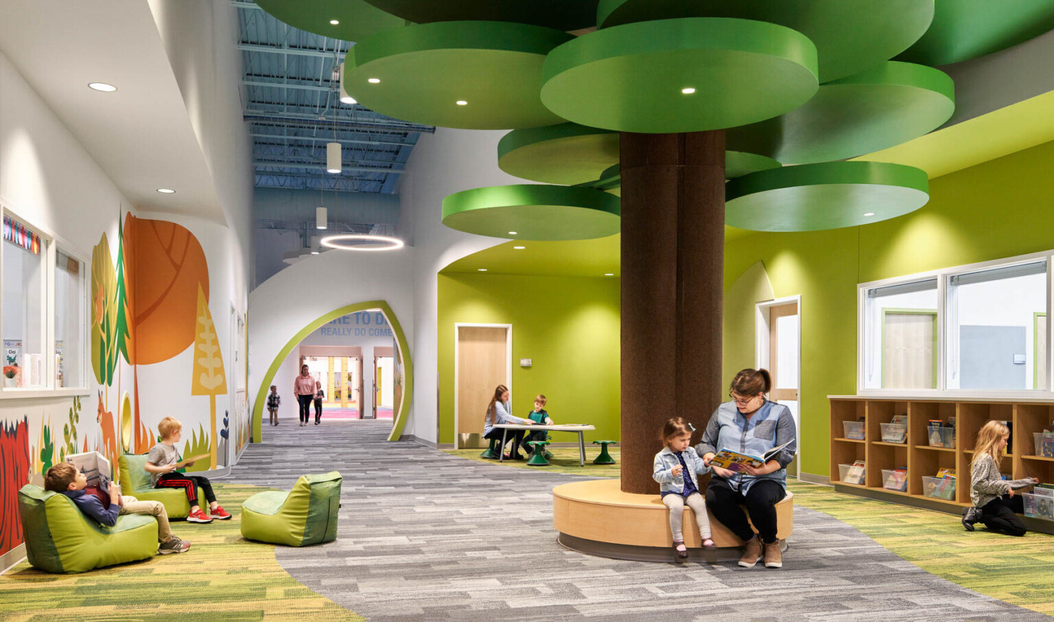people reading on a brown circular bench with a brown column and green ceiling fixtures in hallway lined with forest graphics people reading on a brown circular bench with a brown column and green ceiling fixtures in hallway lined with forest graphics