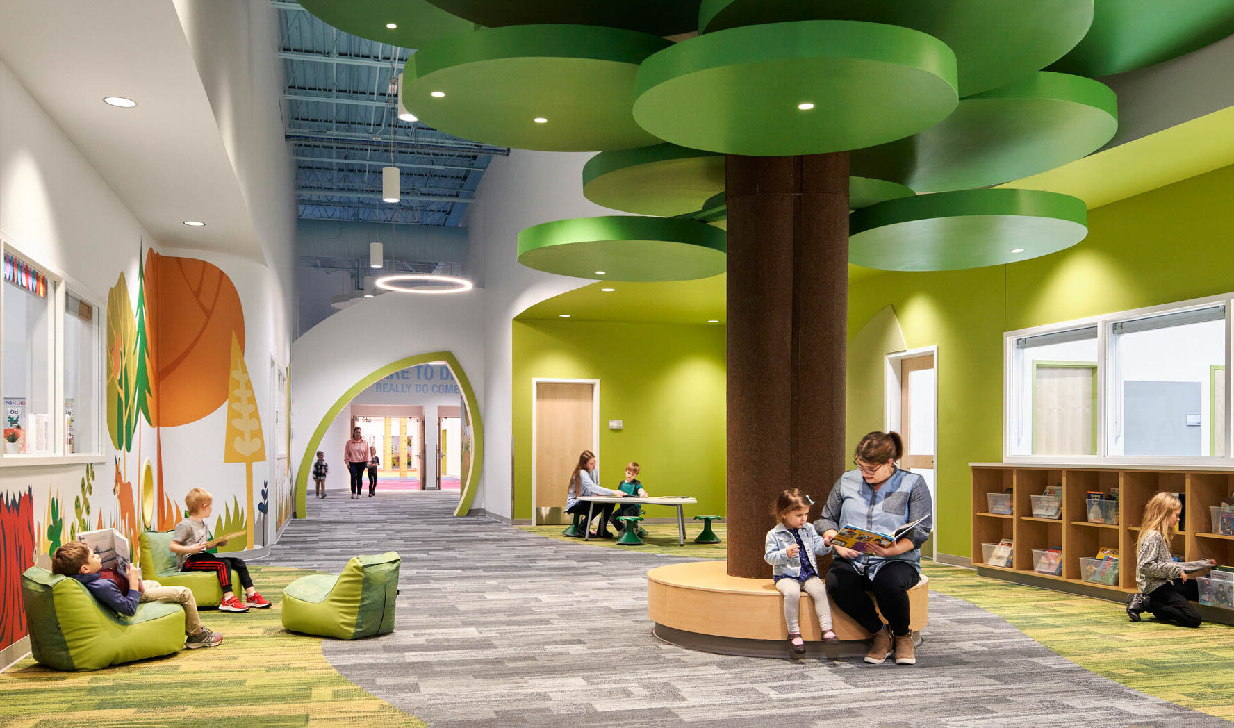 people reading on a brown circular bench with a brown column and green ceiling fixtures in hallway lined with forest graphics