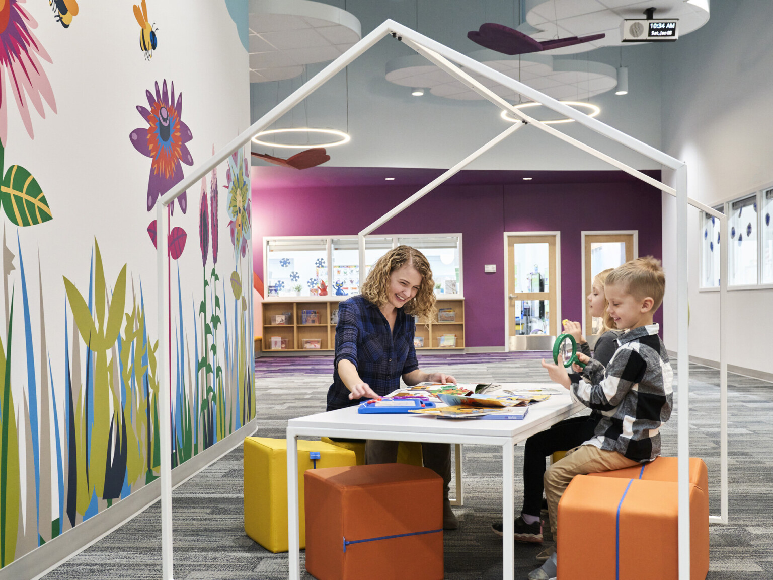 Educator and students reading on flexible comfortable yellow stools at white table with house shaped frame, bright graphic mural Educator and students reading on flexible comfortable yellow stools at white table with house shaped frame, bright graphic mural