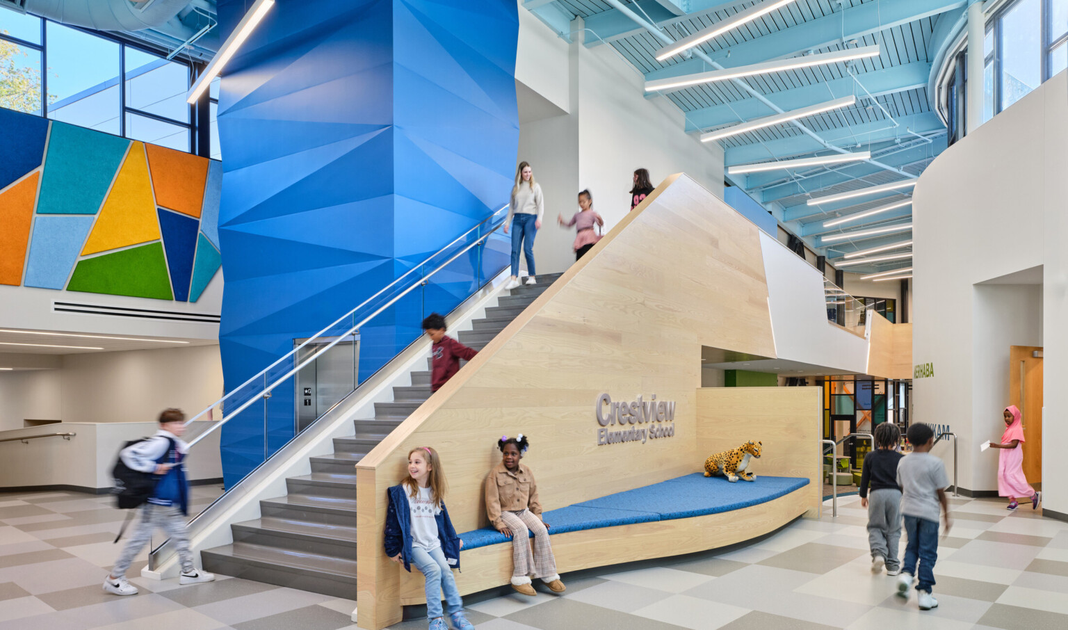Students at Crestview Elementary School in a major hallway with staircase to second floor; blue centerwall divides the floor; Double height overlook is multicolored shape collage